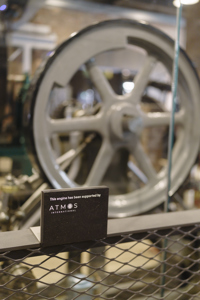 Close-up of the restored Diagonal Steam Engine at the Science and Industry Museum’s Power Hall. A black plaque in front reads: “This engine has been supported by Atmos International.” Behind it, the engine’s large grey flywheel and metal components are visible. Science Museum Group © The Board of Trustees of the Science Museum
