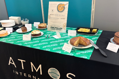 A range of different cakes with corresponding name cards on an Atmos-branded table with a Macmillan tablecloth and a coffee morning poster.