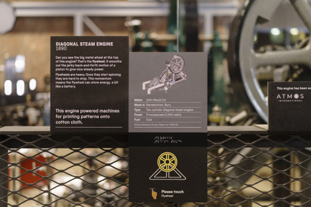 Interpretive panel for the 1890 Diagonal Steam Engine at the Science and Industry Museum’s Power Hall, with an Atmos International support plaque beside it. The text explains that the engine powered machines for printing patterns onto cotton cloth and lists: “Maker: John Wood Ltd, Made in Ramsbottom, Bury, Type: Two cylinder diagonal steam engine, Power: 5 horsepower / 3,500 watts, Fuel: Coal.” A sign below invites visitors to “Please touch Flywheel.” Science Museum Group © The Board of Trustees of the Science Museum